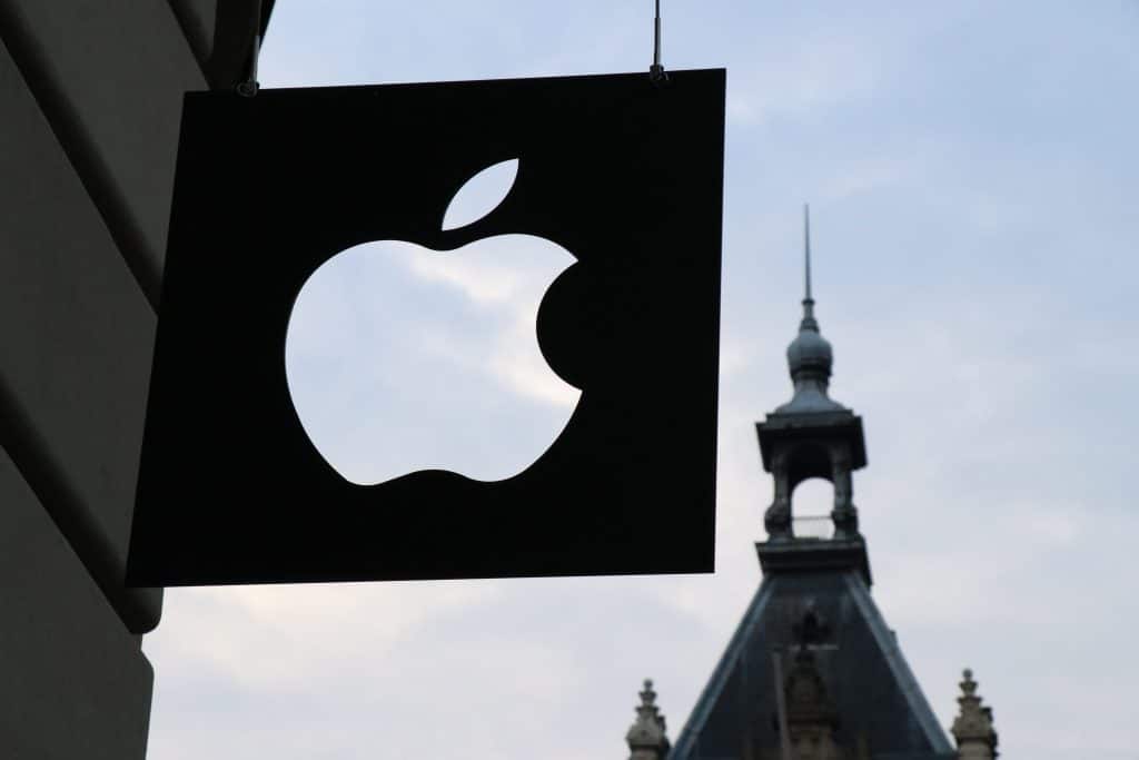 A black sign with a white Apple logo hangs outside a building against a backdrop of a historic structure with a pointed tower, securing its presence even on an overcast sky.