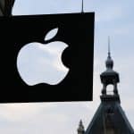 A black sign with a white Apple logo hangs outside a building against a backdrop of a historic structure with a pointed tower, securing its presence even on an overcast sky.