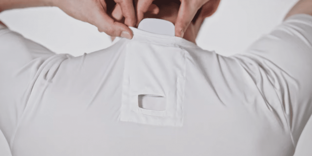 A person wearing a white shirt is adjusting a small, flat white device attached to the back of their neck. The device, part of Sony's personal air conditioning system, is tucked into a pocket with a square cut-out on the shirt's neck area, showcasing a modern design against a plain white background.