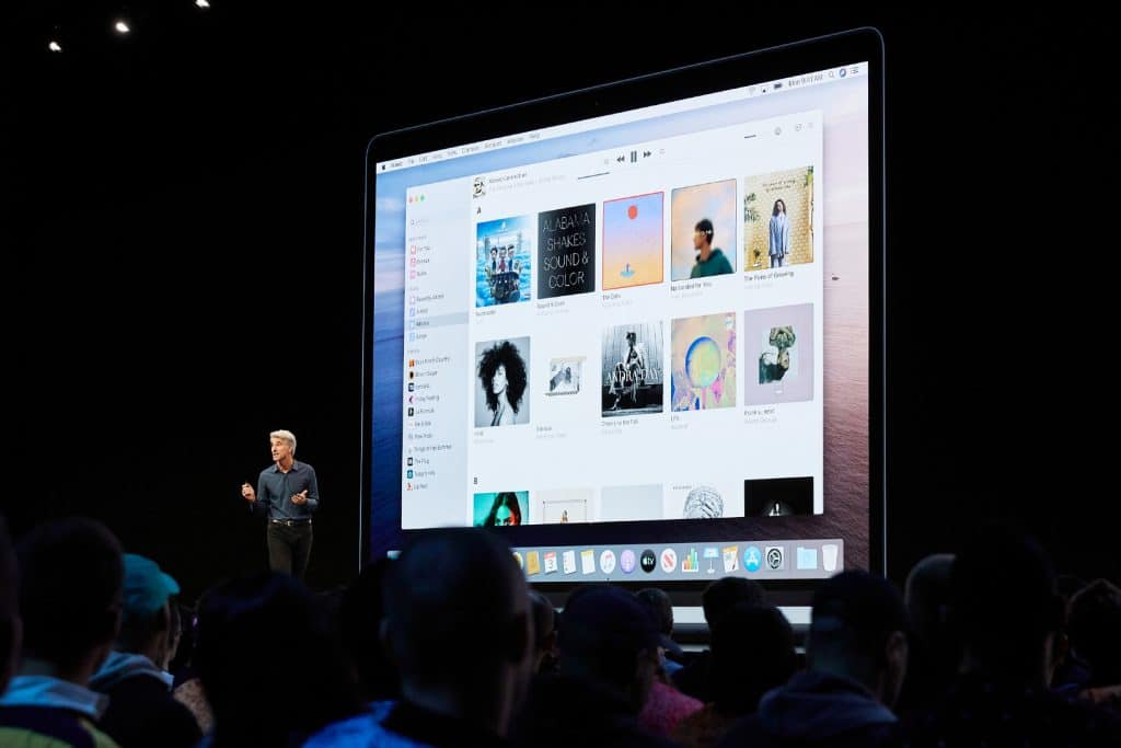 A person is presenting on stage in front of a large audience, looking forward with enthusiasm. The background features a giant screen displaying a music app interface with album covers, similar to the updated look introduced in macOS Catalina. The interface shows different music categories and some visible album names and artwork.