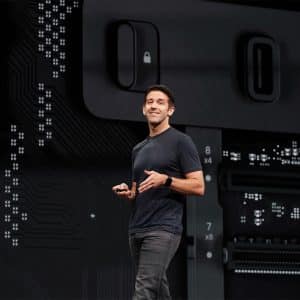 A person in a dark t-shirt and jeans stands on a stage, smiling and holding a device. Behind them, an enlarged image of a tech circuit board showcases various electronic components and connectors, reminiscent of everything we know about the Mac Pro’s advanced technology.