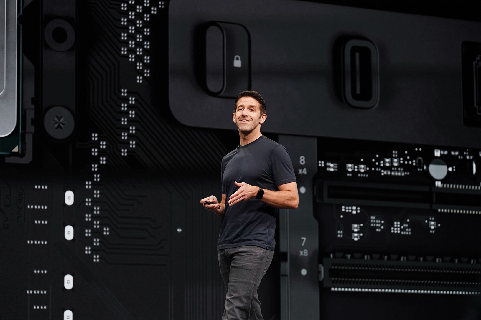 A person in a dark t-shirt and jeans stands on a stage, smiling and holding a device. Behind them, an enlarged image of a tech circuit board showcases various electronic components and connectors, reminiscent of everything we know about the Mac Pro’s advanced technology.