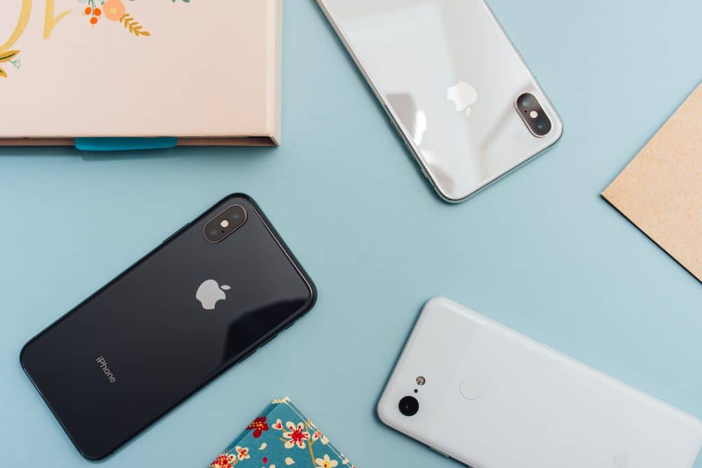 Image shows a flat lay of three smartphones on a light blue surface. There are two iPhones, one black and one silver, highlighting iPhone loyalty even at its lowest levels, along with a white Google Pixel. Additionally, there are partial views of two flowered items and a beige box in the corners of the image.