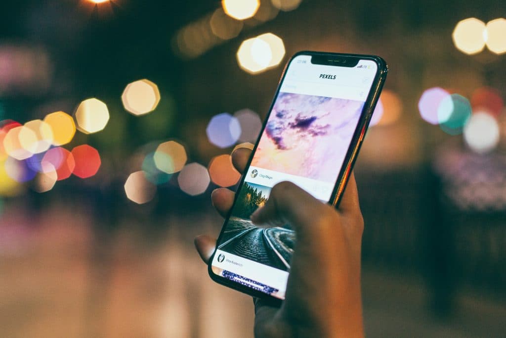 A hand is holding a smartphone, displaying a social media feed with photos of a sunset and train tracks. The background is blurred, with colorful bokeh lights creating a vibrant cityscape at night. The latest iOS 13 beta highlights subtle changes that enhance the overall user experience.