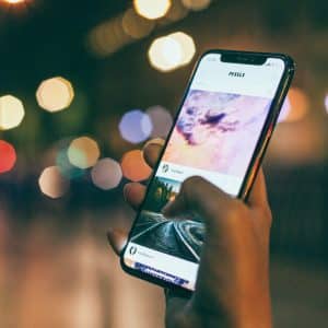 A hand is holding a smartphone, displaying a social media feed with photos of a sunset and train tracks. The background is blurred, with colorful bokeh lights creating a vibrant cityscape at night. The latest iOS 13 beta highlights subtle changes that enhance the overall user experience.