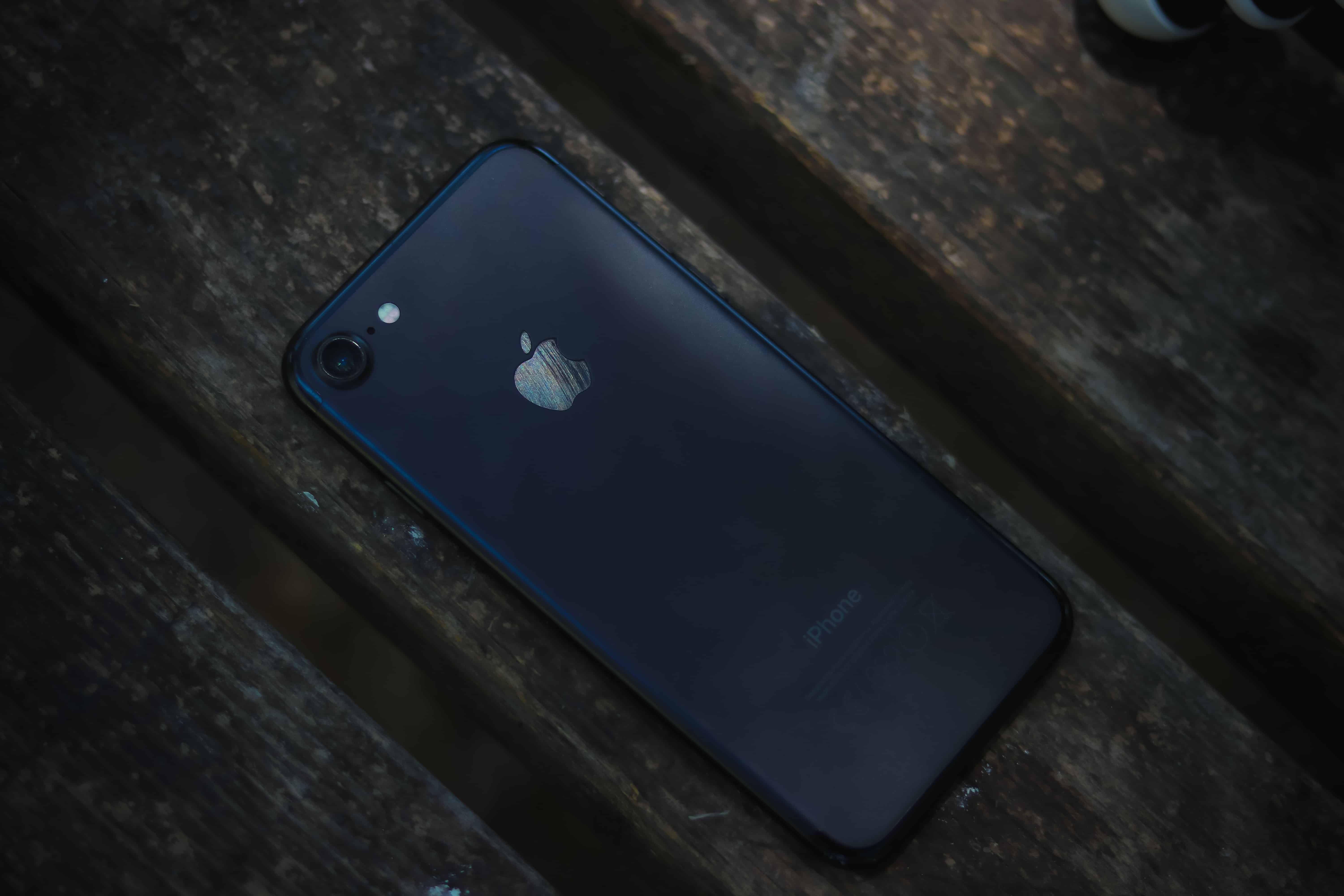 A black iPhone is placed face down on a wooden surface, with the Apple logo clearly visible. The lighting is dim, casting shadows on the phone and the wooden background, reminiscent of minimalist designs often seen in India.