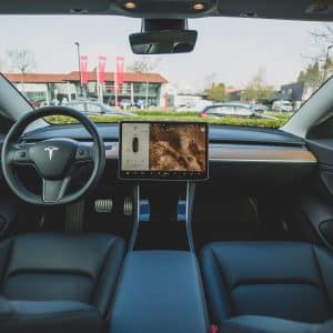 View of a Tesla car's interior showcasing a minimalist dashboard. The center console features a large touchscreen display with navigation and media controls. Black seats and modern design elements are highlighted, while buildings and cars are visible through the windshield, resonating subtly with Apple’s sleek design ethos.