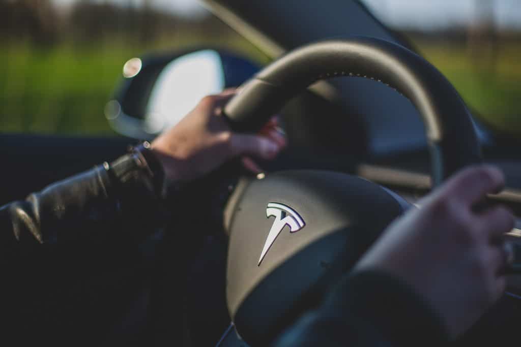 Close-up of a Tesla engineer driving a Tesla car, both hands gripping the steering wheel adorned with the iconic Tesla logo. The background is blurred, offering a glimpse of greenery and the rearview mirror on the left side.