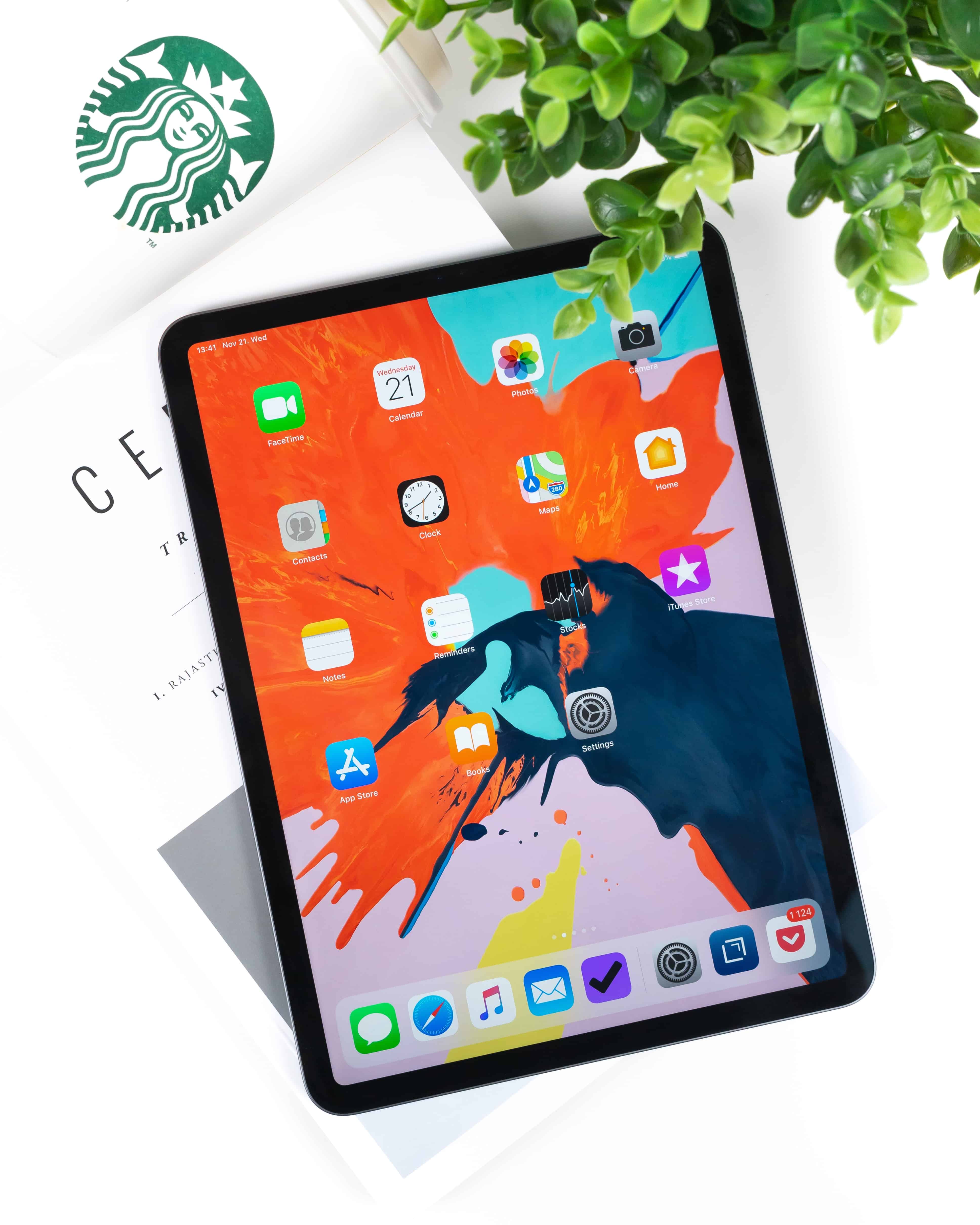 An iPad displaying a colorful home screen with various iPadOS apps, partially covering an open book. A Starbucks logo is visible in the upper left corner, and a leafy green plant is positioned in the top right, exemplifying Project Catalyst's seamless integration of mobile and desktop environments.