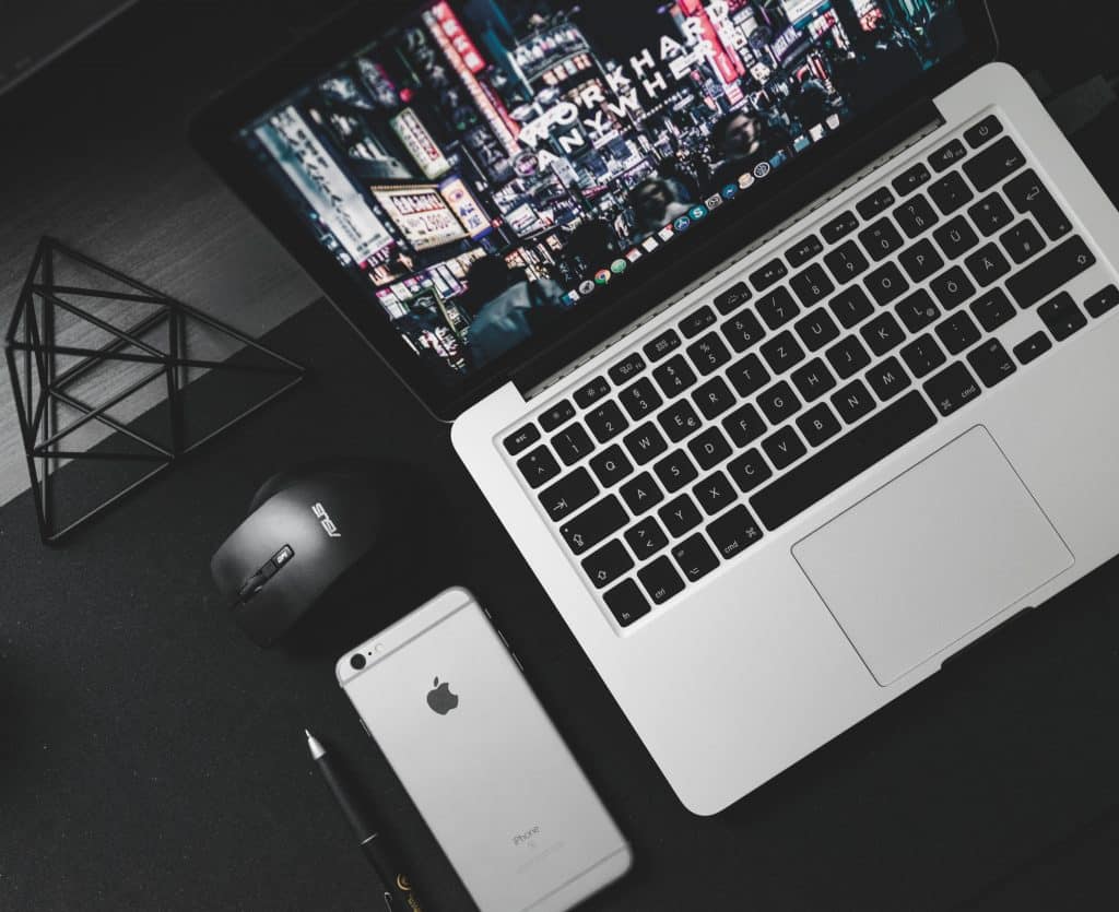 A silver MacBook with a butterfly keyboard and cityscape screensaver sits on a black desk next to a black ASUS computer mouse and a silver iPhone. A black geometric desk decoration and a pen are also present. The dark desk surface creates a sleek, minimalist aesthetic, perfect for new Macs.