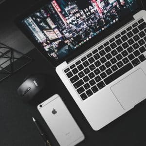 A silver MacBook with a butterfly keyboard and cityscape screensaver sits on a black desk next to a black ASUS computer mouse and a silver iPhone. A black geometric desk decoration and a pen are also present. The dark desk surface creates a sleek, minimalist aesthetic, perfect for new Macs.