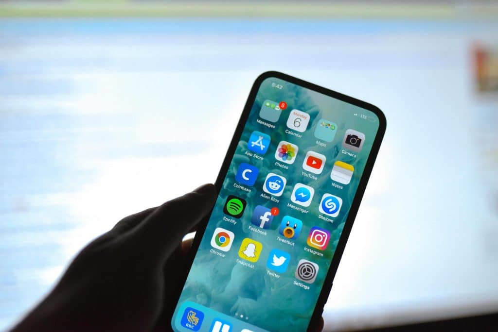 A person holds a notch-less iPhone displaying various app icons, including Messages, Calendar, Photos, YouTube, Facebook, Instagram, Spotify, Snapchat, Twitter, and others. The background shows a blurred computer screen. The person’s hand is slightly shadowed.