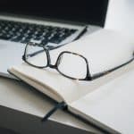 A pair of Apple Glasses rests on an open notebook with lined pages. A black pen lies next to the notebook, and a laptop can be seen in the background. The scene is set on a white desk, suggesting a workspace or study area.