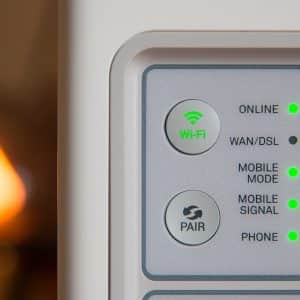 Close-up of a Wi-Fi router displaying various status indicators. The online, WAN/DSL, Wi-Fi, mobile mode, mobile signal, and phone indicators are all illuminated in green, suggesting that the router is functioning correctly. A soft, warm light is blurred in the background. It supports Apple's new WiFi band.