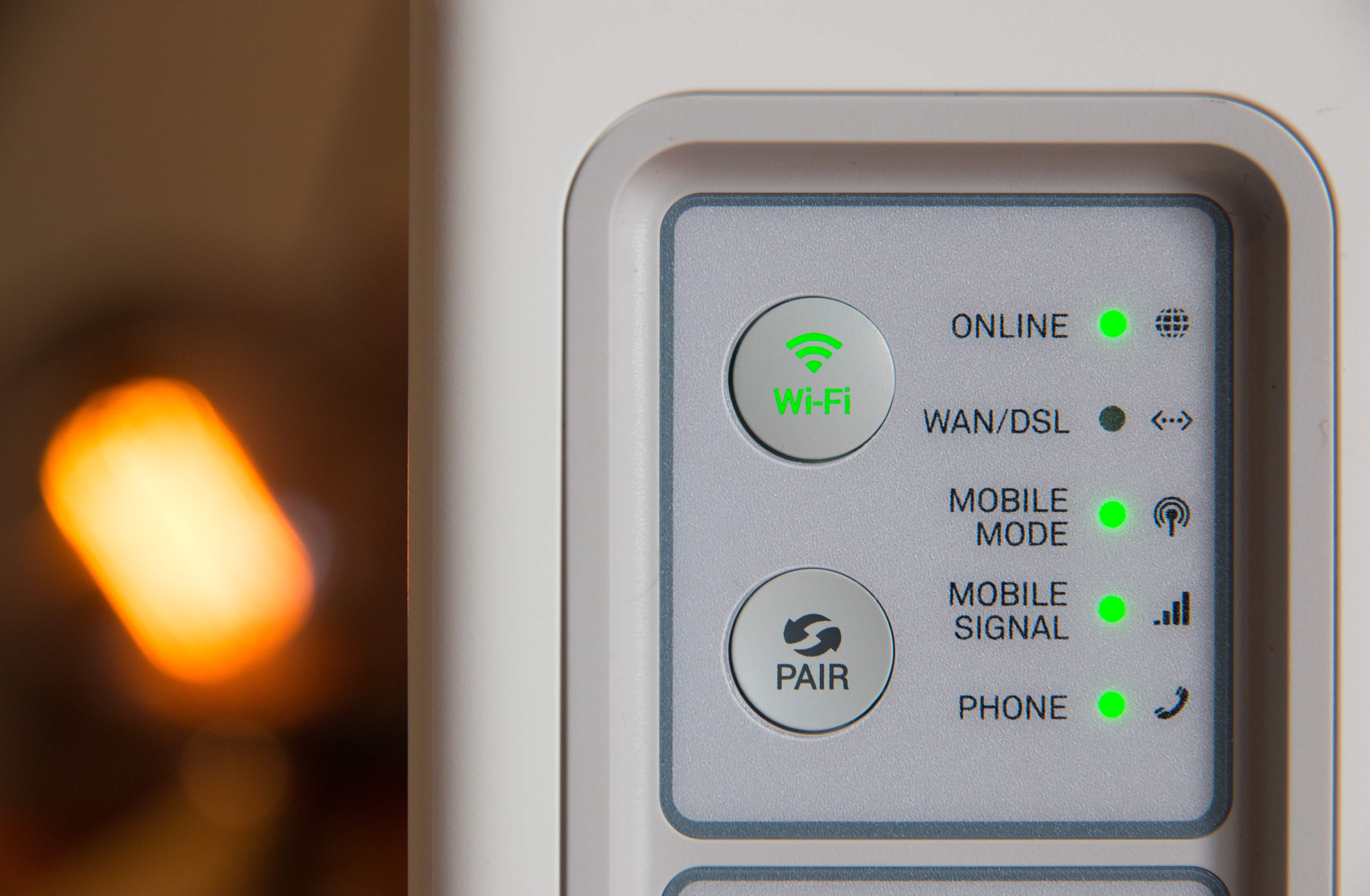 Close-up of a Wi-Fi router displaying various status indicators. The online, WAN/DSL, Wi-Fi, mobile mode, mobile signal, and phone indicators are all illuminated in green, suggesting that the router is functioning correctly. A soft, warm light is blurred in the background. It supports Apple's new WiFi band.