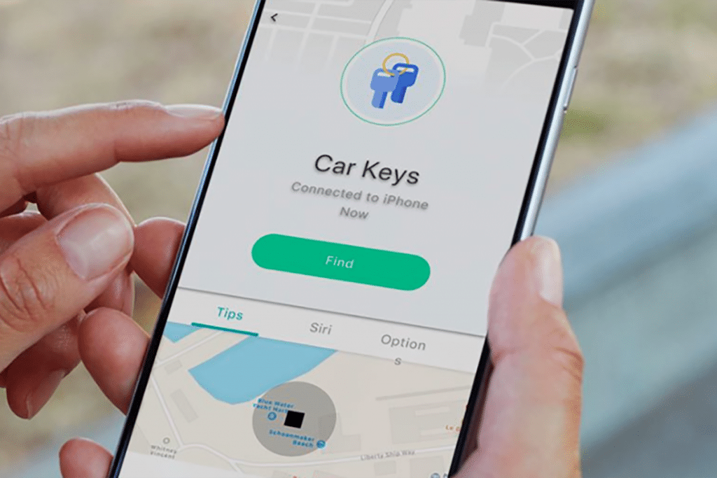 A person holds a smartphone displaying an app interface labeled "Car Keys," connected to the iPhone via Bluetooth beacon. The screen features buttons such as "Find," "Tips," "Siri," and "Option." A partial map is visible at the bottom, showcasing Apple's sleek product design.