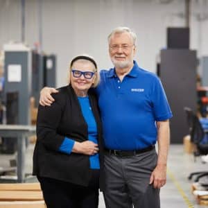 An older man and woman stand in a warehouse-like environment with their arms around each other, smiling. The man wears a blue polo shirt, and the woman sports a black blazer with a blue top and glasses. Proudly responsible for creating 2.4 million jobs, they celebrate their success surrounded by shelves and boxes.