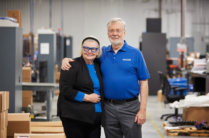 An older man and woman stand in a warehouse-like environment with their arms around each other, smiling. The man wears a blue polo shirt, and the woman sports a black blazer with a blue top and glasses. Proudly responsible for creating 2.4 million jobs, they celebrate their success surrounded by shelves and boxes.