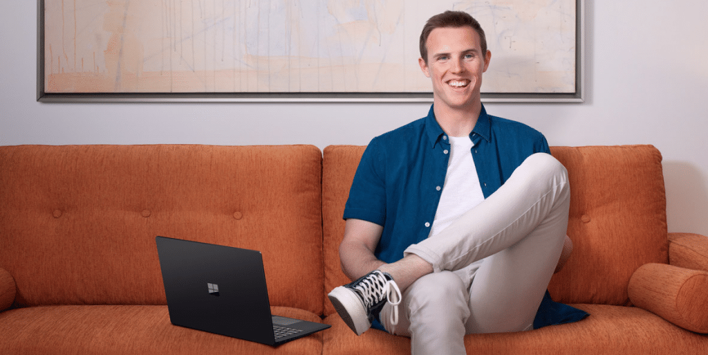 A person with short hair, wearing a blue shirt and white pants, smiles while sitting on an orange couch with one leg crossed over the other. Beside them rests a sleek Microsoft Surface. A minimalistic painting hangs on the wall in the background, hinting at simplicity and style in this new advert.