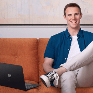 A person with short hair, wearing a blue shirt and white pants, smiles while sitting on an orange couch with one leg crossed over the other. Beside them rests a sleek Microsoft Surface. A minimalistic painting hangs on the wall in the background, hinting at simplicity and style in this new advert.