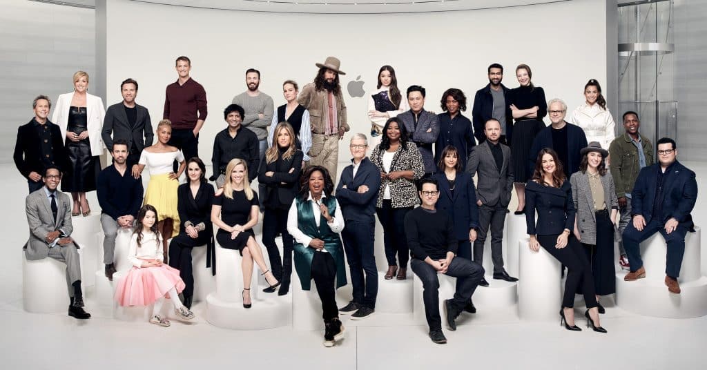 A group of 28 people, including men and women of various ages, dressed in a mix of formal and casual attire. They are arranged in rows, some sitting and others standing, against a simple, light-colored background with cylindrical platforms. It's as organized as simultaneous streams on Apple TV+.