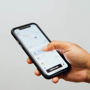 A person holds an Apple iPhone displaying a ride-sharing app with a map and car options on the screen. The person's thumb is poised to select one of the ride options. The background is plain and light-colored.