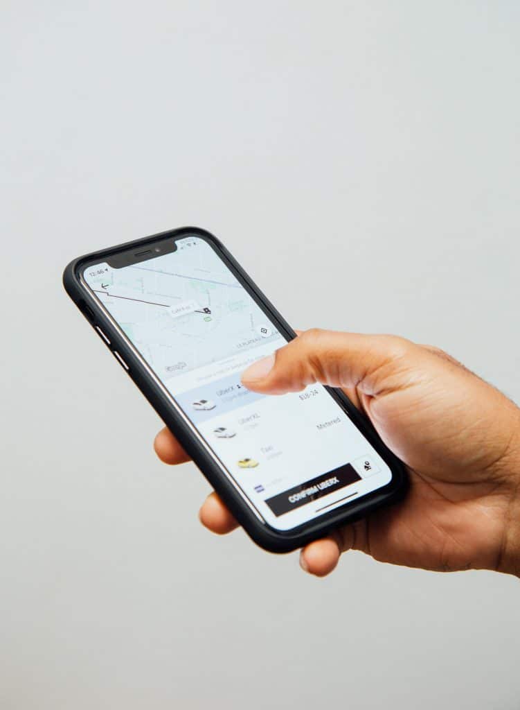 A person holds an Apple iPhone displaying a ride-sharing app with a map and car options on the screen. The person's thumb is poised to select one of the ride options. The background is plain and light-colored.