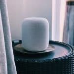 A white cylindrical smart speaker with a mesh exterior sits on a round, woven black table. This 2020 release from Apple resembles a Cheaper HomePod. The speaker is placed on a metallic tray, and a curtain is partially visible to the left, with blurred background elements on the right.