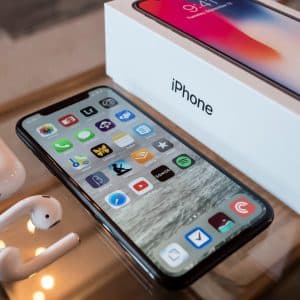 A new iPhone with its screen on sits next to its white packaging box, a pair of wireless earpods, and a charging case on a reflective surface. The iPhone displays various app icons along with an updating warning message, while the box features the "iPhone" label.