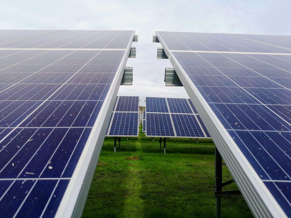 Close-up view of solar panels aligned in rows on a green grassy field in Taiwan, with a cloudy sky in the background. The image captures the perspective between two rows of panels, emphasizing solar energy as a clean, renewable source.