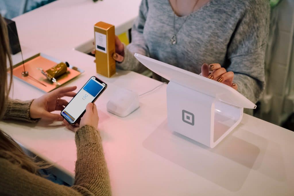 A customer is using their smartphone with Apple Pay to make a payment through a white Square terminal at a US retail counter. An employee, partially visible, is assisting with the transaction. A small box and a card reader are on the counter.