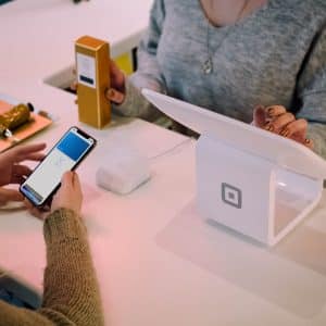 A customer is using their smartphone with Apple Pay to make a payment through a white Square terminal at a US retail counter. An employee, partially visible, is assisting with the transaction. A small box and a card reader are on the counter.