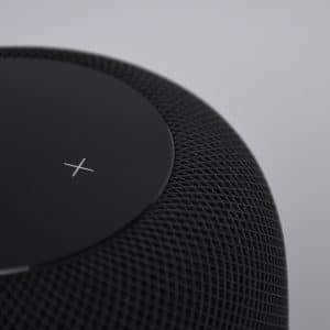 Close-up of the top part of a smart speaker with a mesh exterior. The touch surface on top displays a simple white "X" on a black background. The smart speaker, resembling the sleek design of a HomePod, is set against a plain, light-colored background.