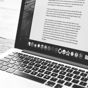 A grayscale photo of an open Apple laptop on a wooden surface, displaying a document filled with text. The laptop has a sleek design, and various application icons are visible in the dock at the bottom of the screen. The overall scene appears minimalistic and modern, subtly hinting at Apple's commitment to tackling security issues through their bug bounty program.