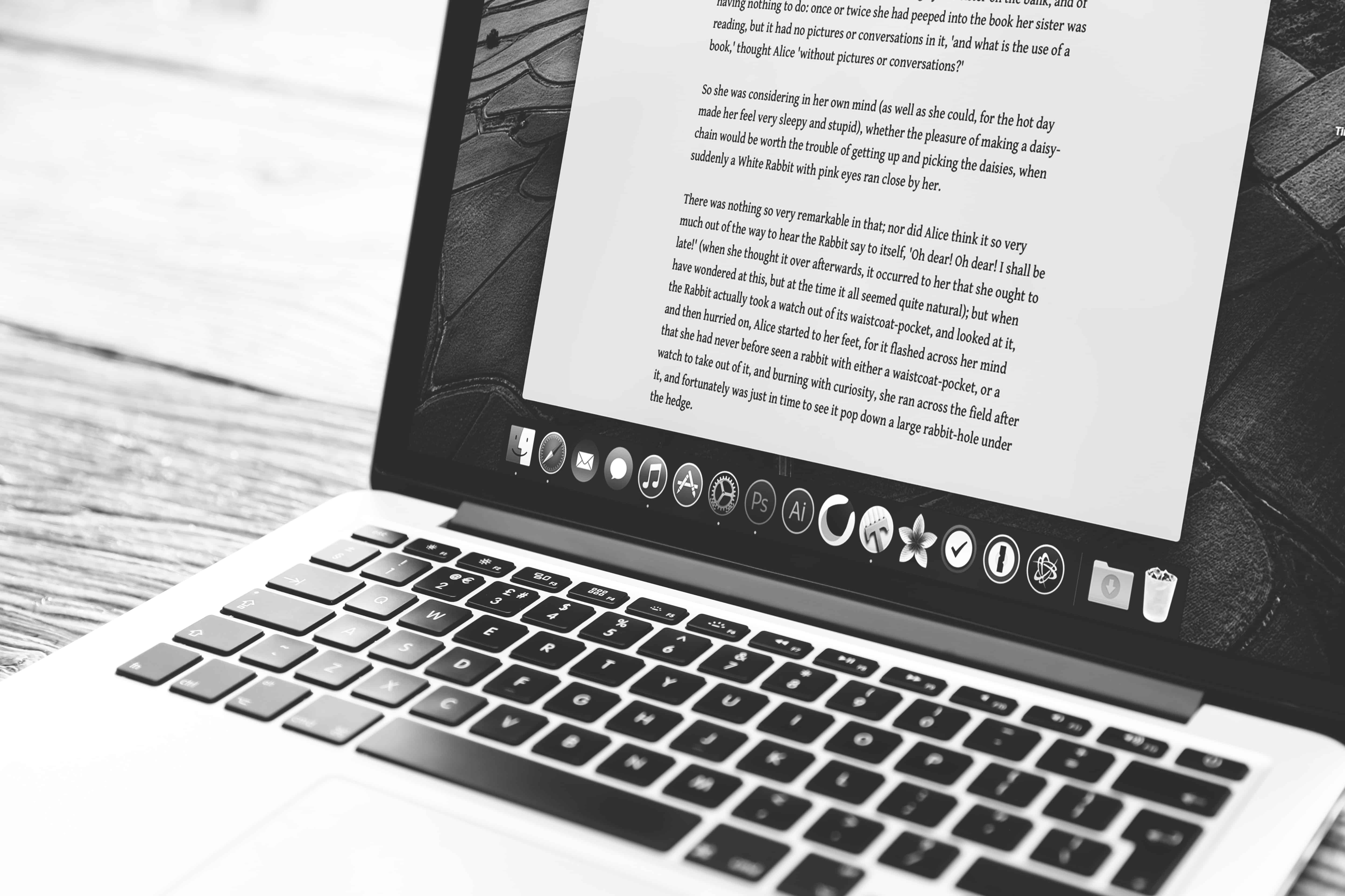 A grayscale photo of an open Apple laptop on a wooden surface, displaying a document filled with text. The laptop has a sleek design, and various application icons are visible in the dock at the bottom of the screen. The overall scene appears minimalistic and modern, subtly hinting at Apple's commitment to tackling security issues through their bug bounty program.