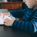 A child, dressed in a blue and black striped shirt, focuses on a tablet screen while seated at a wooden table. The child is using both hands to interact with kids apps on the device. In the background, blurred furniture creates a cozy atmosphere.