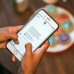 A person holding a smartphone over a wooden table types a message. On the table, there are colorful, decorated cookies in the background. The screen shows a conversation within a messaging app discussing how Google shares details about the largest attack against iPhone users.