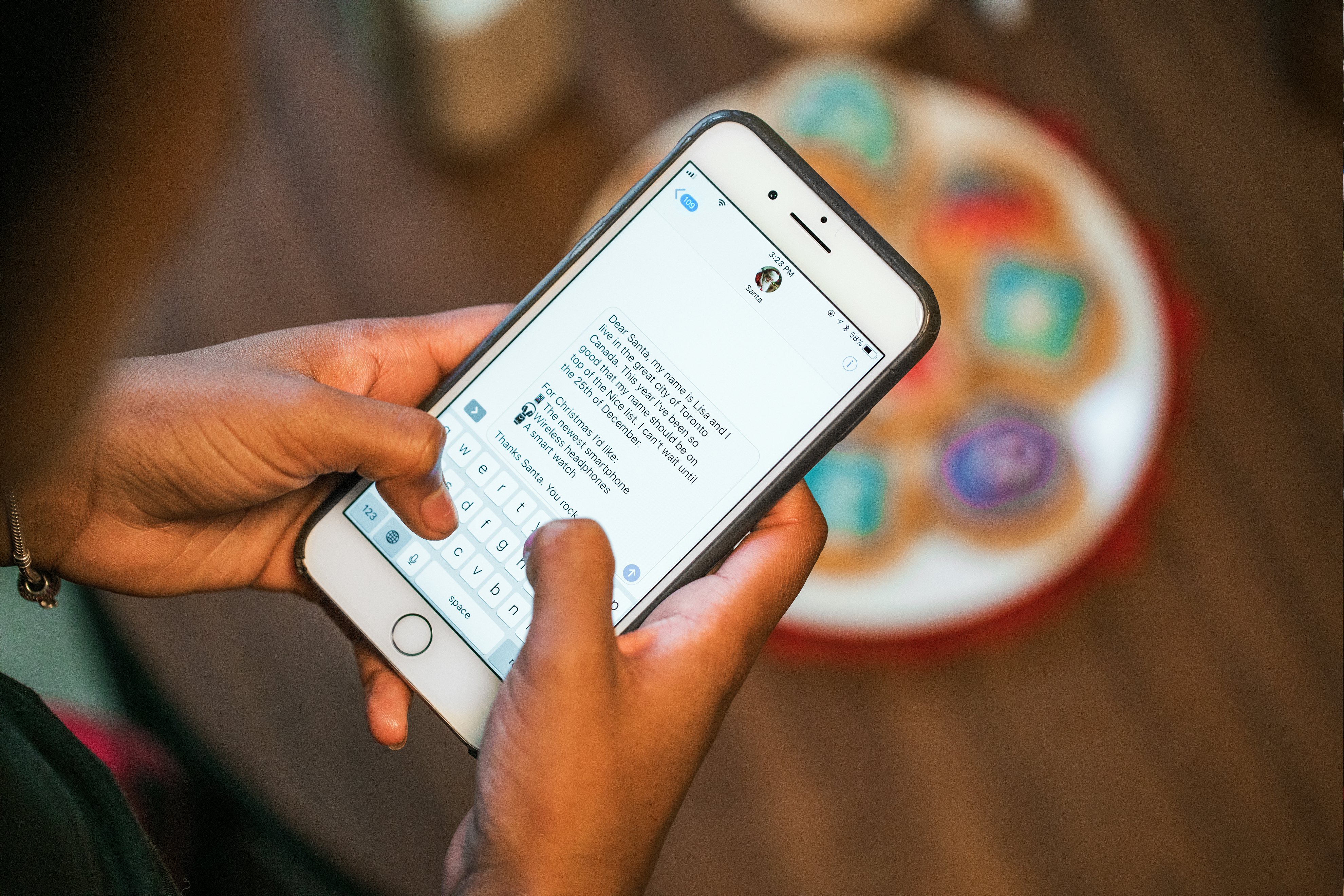 A person holding a smartphone over a wooden table types a message. On the table, there are colorful, decorated cookies in the background. The screen shows a conversation within a messaging app discussing how Google shares details about the largest attack against iPhone users.