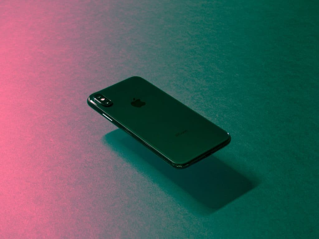 A green iPhone is displayed against a gradient background with shades of pink and green. The Apple logo is visible in the center of the back of the phone. Positioned at a slight angle with a shadow adding depth, this image subtly alludes to Apple's response to the battery locking issue.