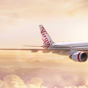 An airplane with the "Virgin Australia" logo on its tail and "virgin" written along its fuselage flies above a layer of clouds during a sunset. The sky is filled with soft hues of pink, orange, and yellow.