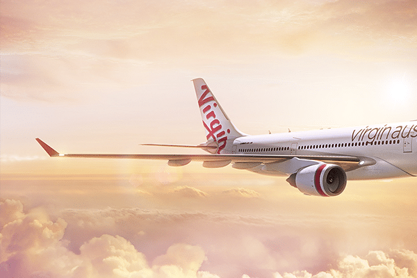 An airplane with the "Virgin Australia" logo on its tail and "virgin" written along its fuselage flies above a layer of clouds during a sunset. The sky is filled with soft hues of pink, orange, and yellow.