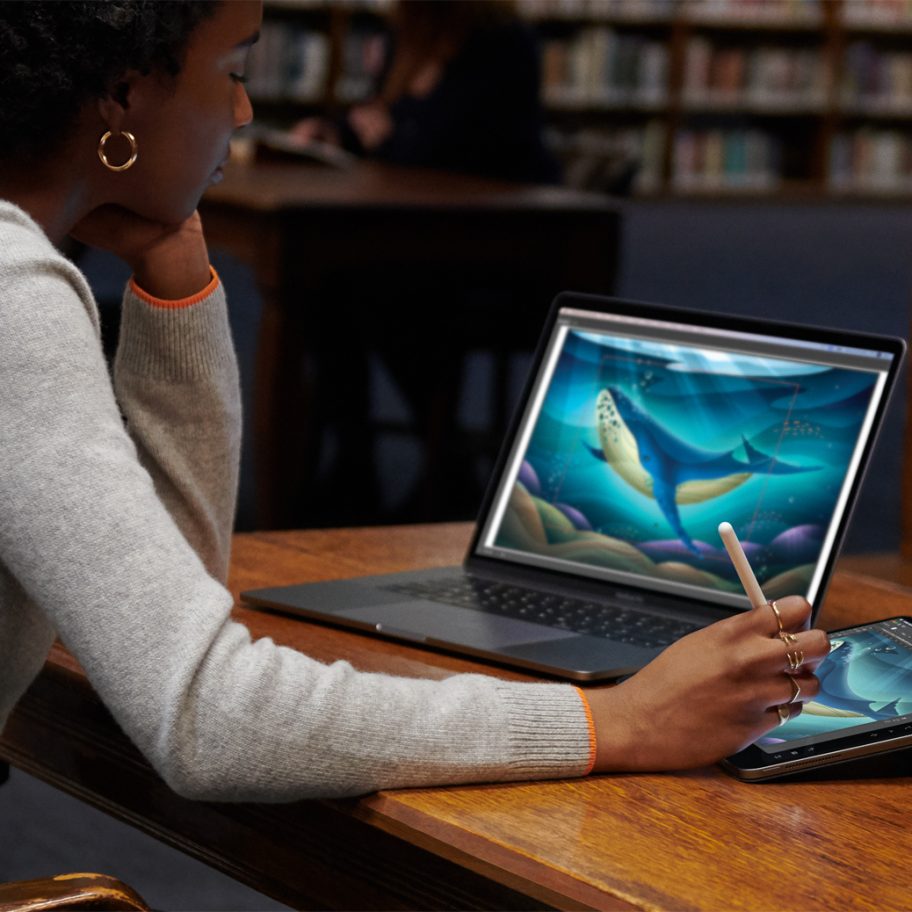 A person uses a tablet with a stylus while also working on a laptop at a desk in a library. Both screens show the same illustration of a seal swimming underwater. Bookshelves filled with books are visible in the background.