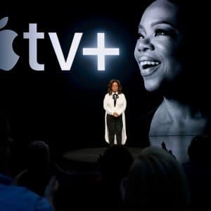 A person stands on stage with a large screen behind them displaying the Apple TV+ logo and a portrait of a smiling person. In their white blouse and black pants, they address the audience about exciting new content launching on November 1, including highlights from Oprah’s Book Club.