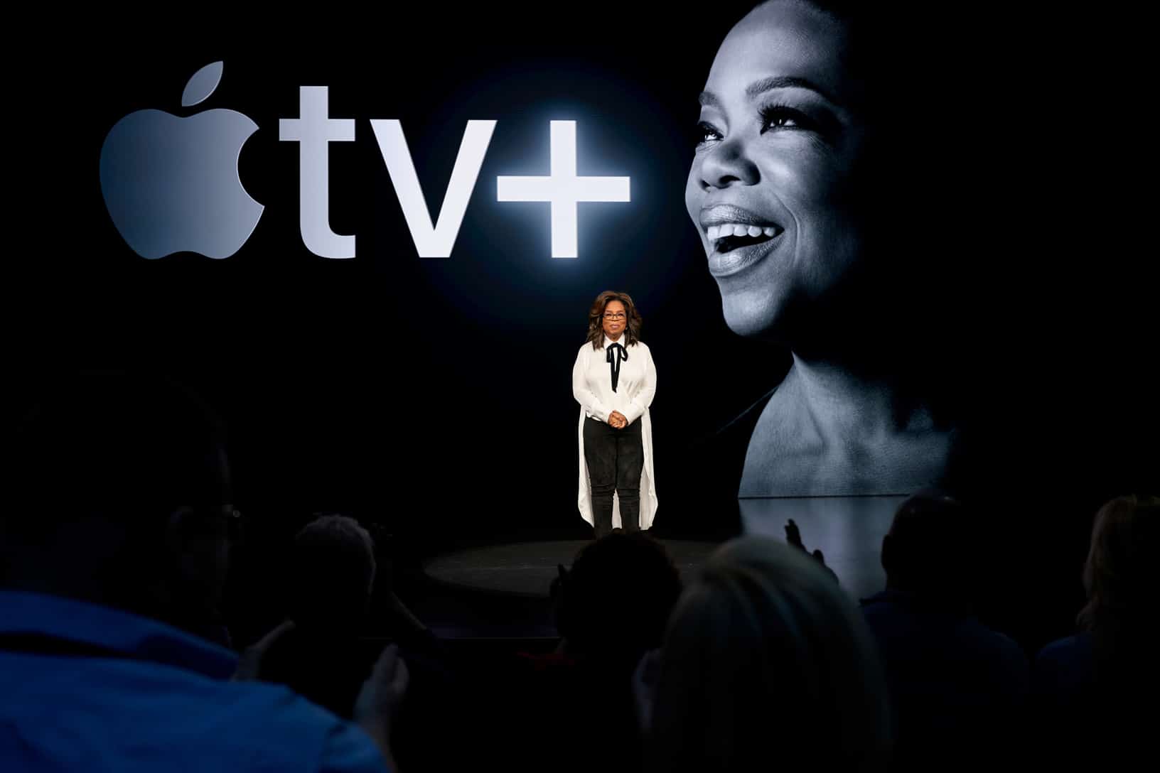 A person stands on stage with a large screen behind them displaying the Apple TV+ logo and a portrait of a smiling person. In their white blouse and black pants, they address the audience about exciting new content launching on November 1, including highlights from Oprah’s Book Club.