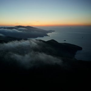 A serene coastal landscape at dusk, featuring rolling hills partially shrouded in mist. The horizon glows with the last light of sunset, casting a warm gradient from orange to deep blue over the calm ocean and distant islands. Perfect for those seeking macOS Catalina wallpapers to enhance their screens.