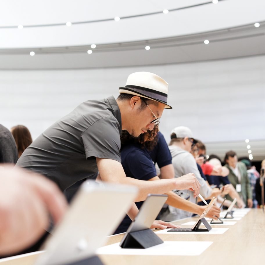 People gather around a large table, interacting with various tablets and devices. A man in a hat closely inspects a tablet, while others in the background are engaged with the devices as well. The room is brightly lit and appears to be a modern, open retail environment.