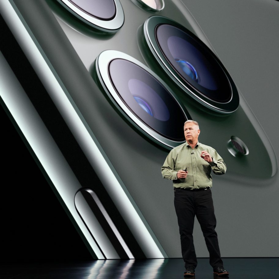 A man in a green shirt and black pants is speaking on a stage. Behind him is a large projection of a smartphone's triple camera module. The image highlights the phone's sleek design and advanced camera technology.