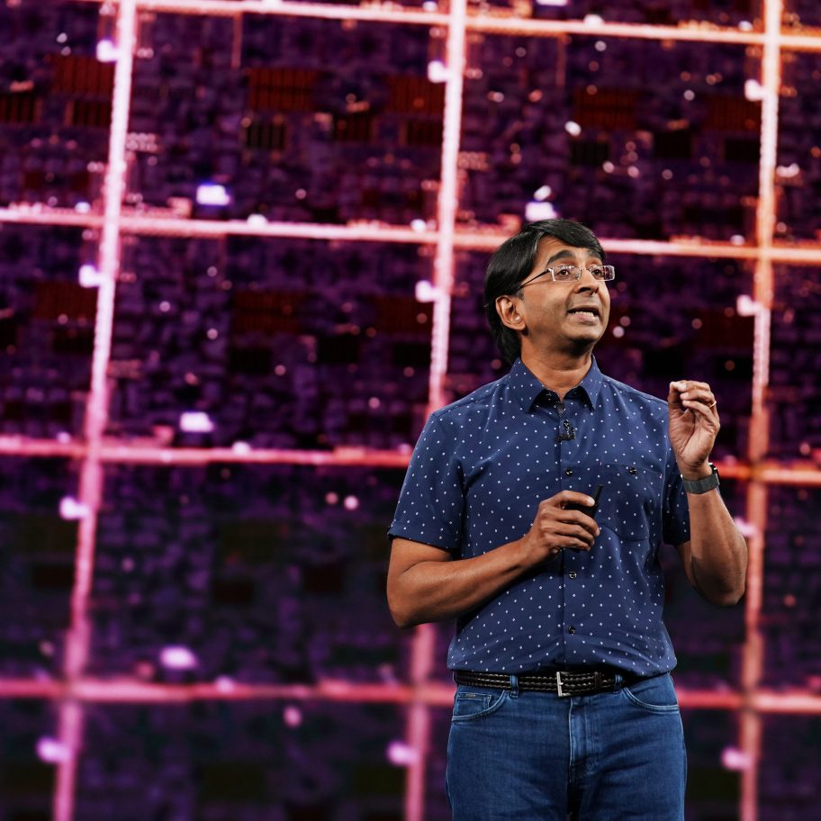 A person wearing glasses and a blue polka dot shirt holds a microphone while speaking on a stage. Behind them is a large screen displaying a close-up, grid-like pattern. The person appears engaged and focused on delivering their presentation.