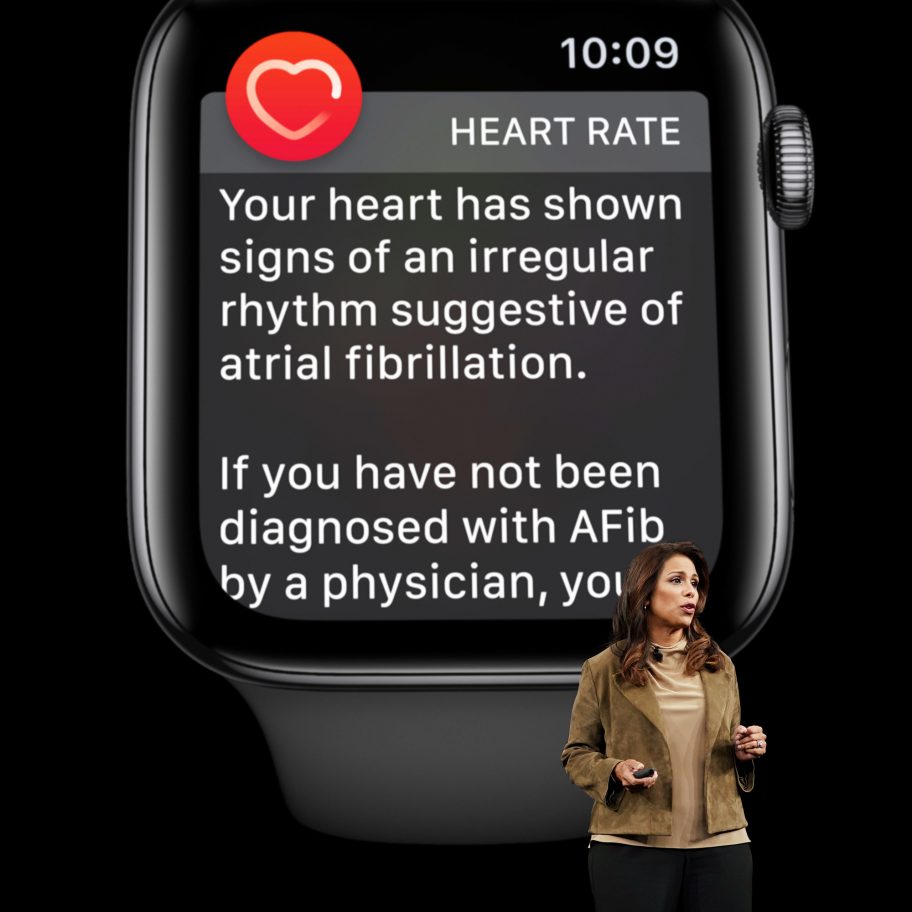 A smartwatch screen displays a heart rate alert, indicating signs of an irregular rhythm suggestive of atrial fibrillation. A person stands in the foreground, pointing to the screen while speaking. The time on the watch reads 10:09.