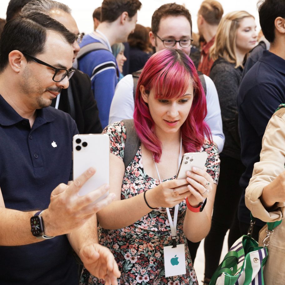 A diverse group of people are gathered closely together, many holding up phones and taking photos or videos. The focus is on a man with glasses and a woman with pink hair, both inspecting their phones. The event seems lively and well-attended.